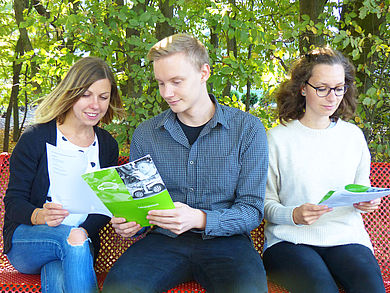 Drei Studenten sitzen auf der Bank und schauen HTW Broschüre an.